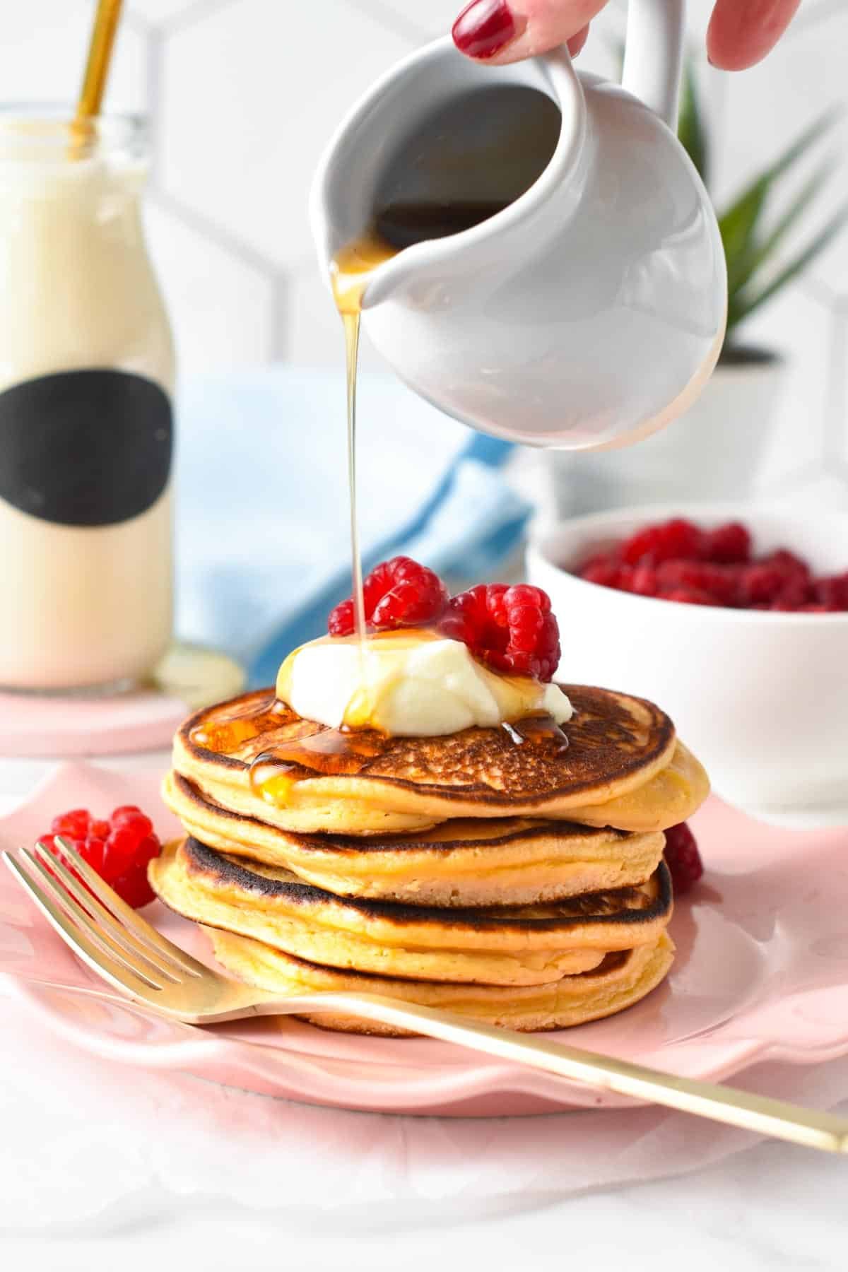 Pouring maple syrup on a stack of 3 ingredient Protein Pancakes decorated with raspberries and Greek yogurt.