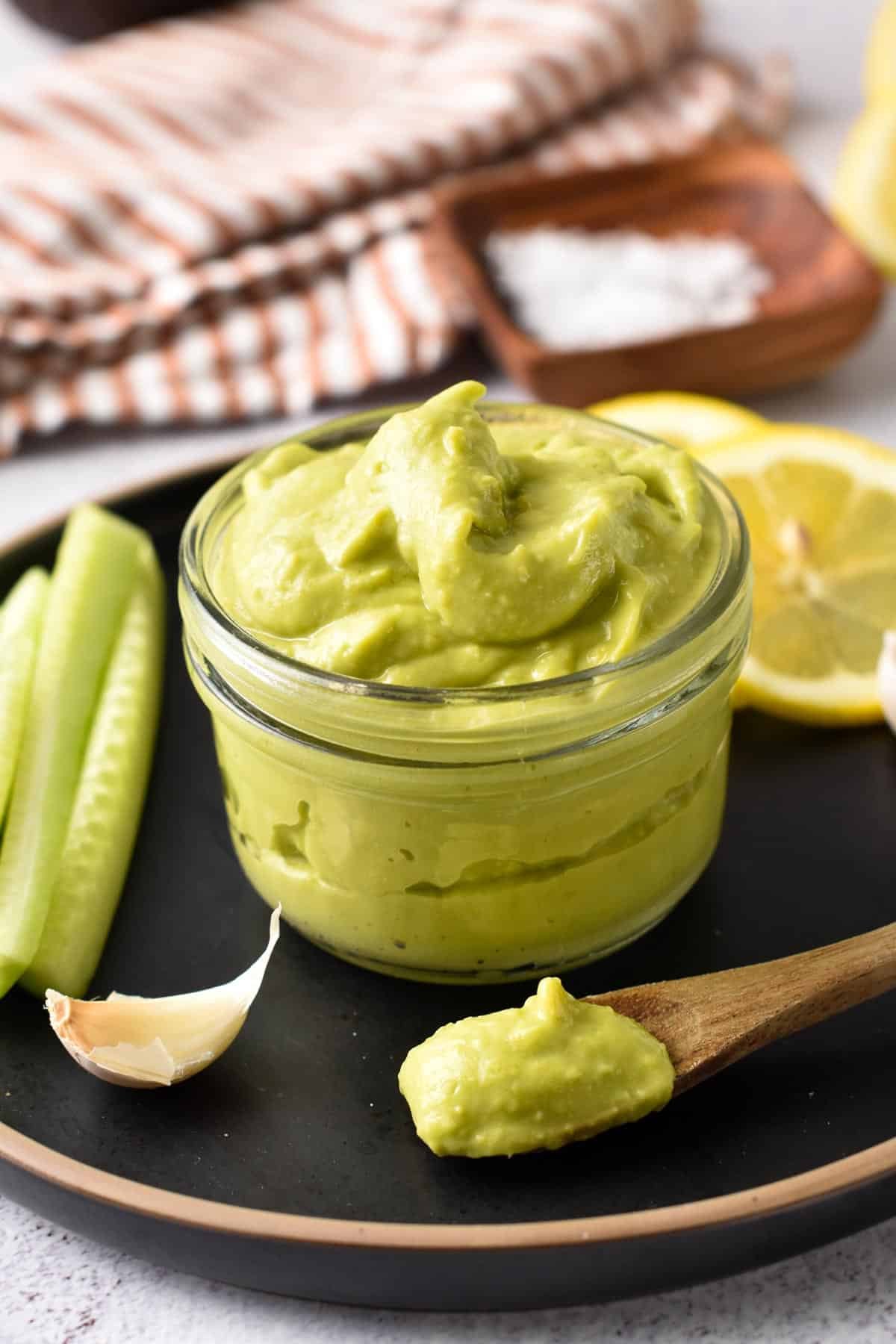 A jar full of avocado mayonnaise on a black plate, in front of lemon slices and cucumber sticks.