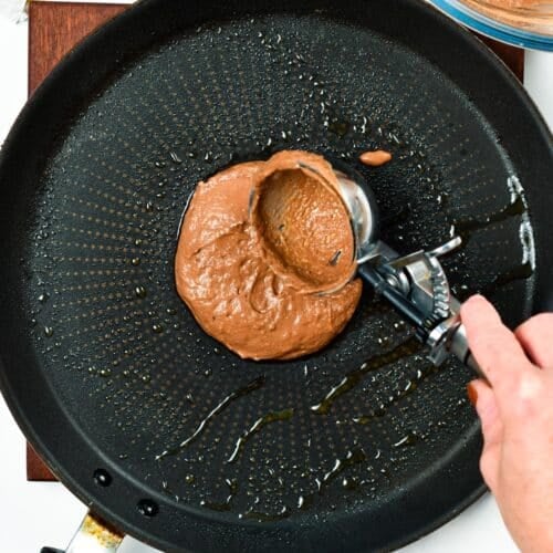 chocolate pancake batter in a warm grease skillet with a cookie dough scoop spreading the batter