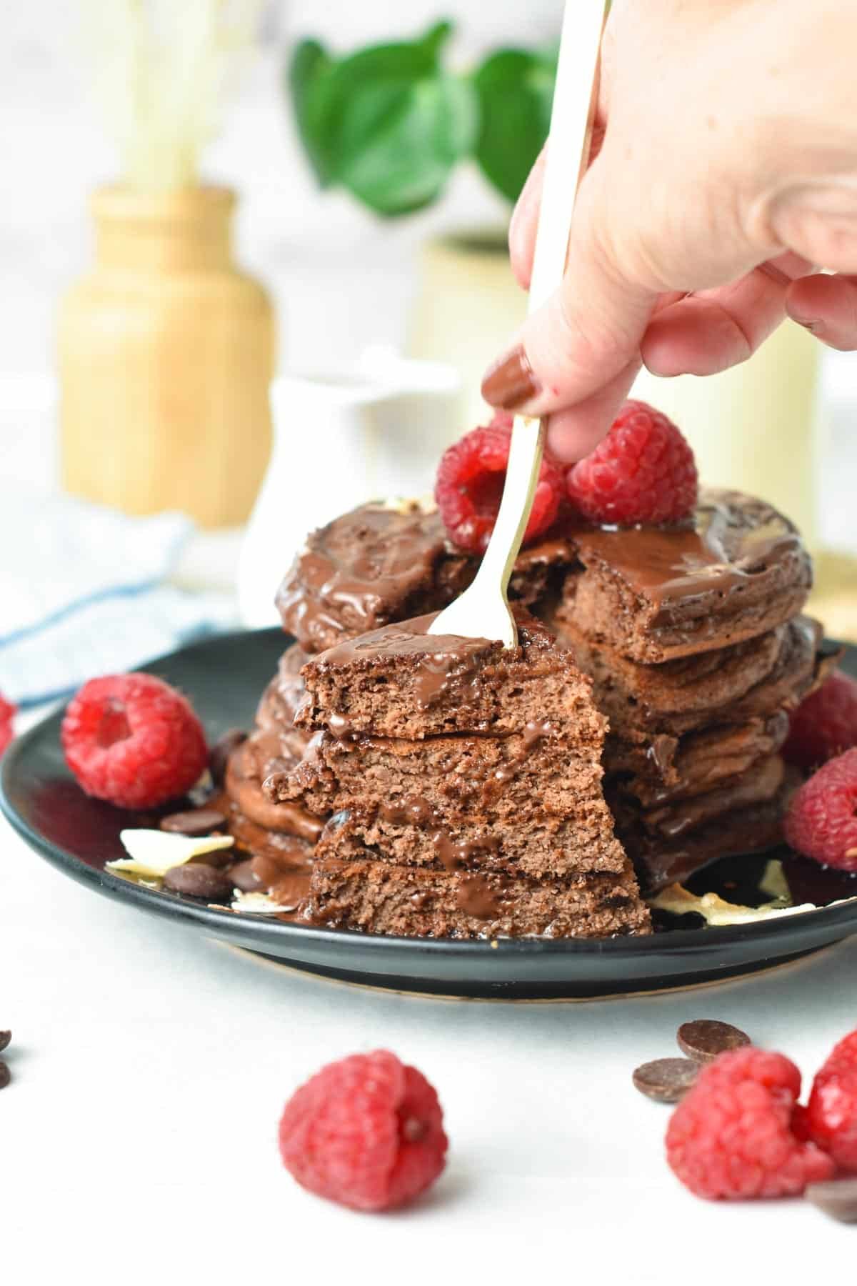 A stack of Chocolate Protein Pancakes with chocolate sauce on top and fresh raspberries and a fork showing a portion of the stack cut.