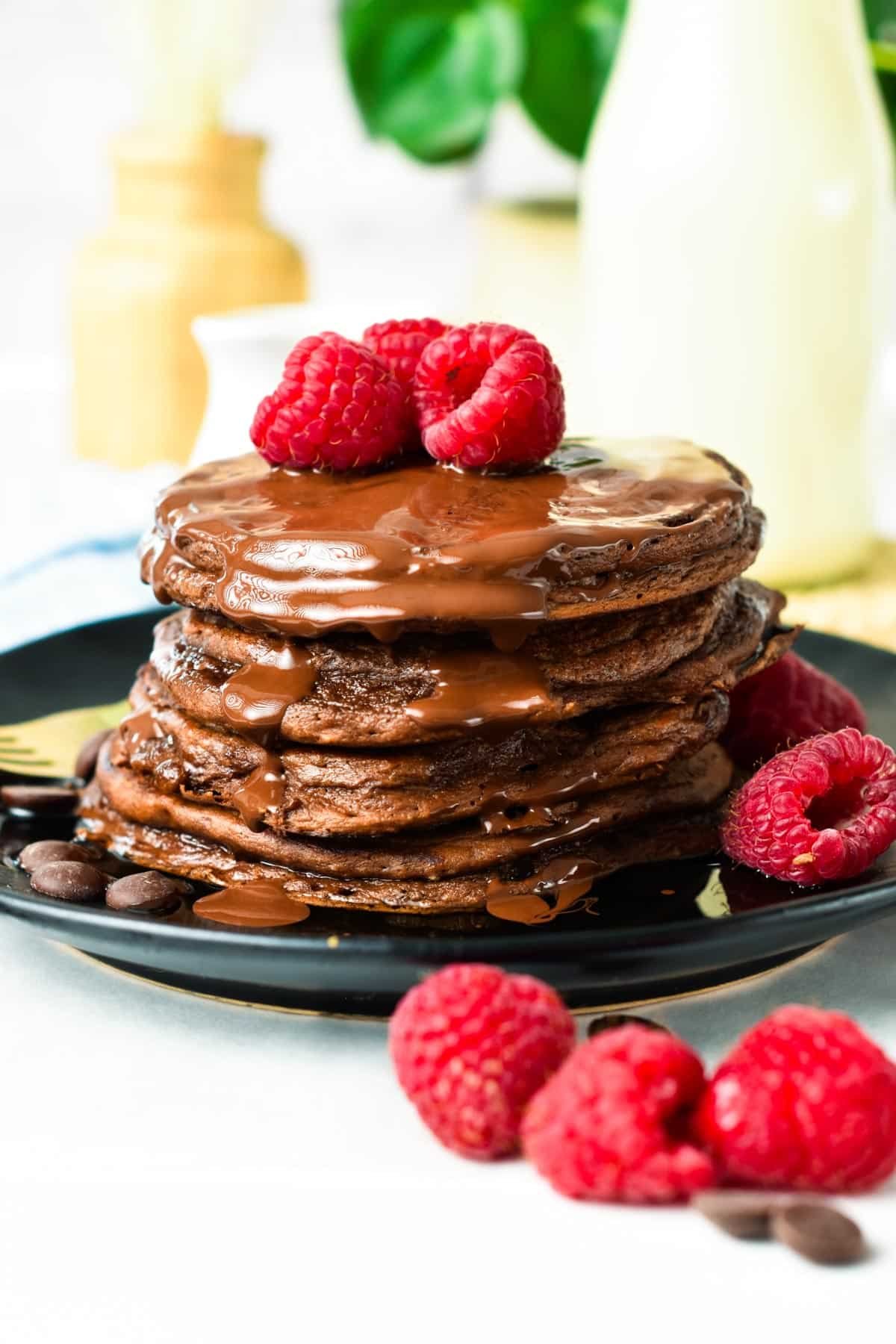 A stack of Chocolate Protein Pancakes with chocolate sauce on top and fresh raspberries.