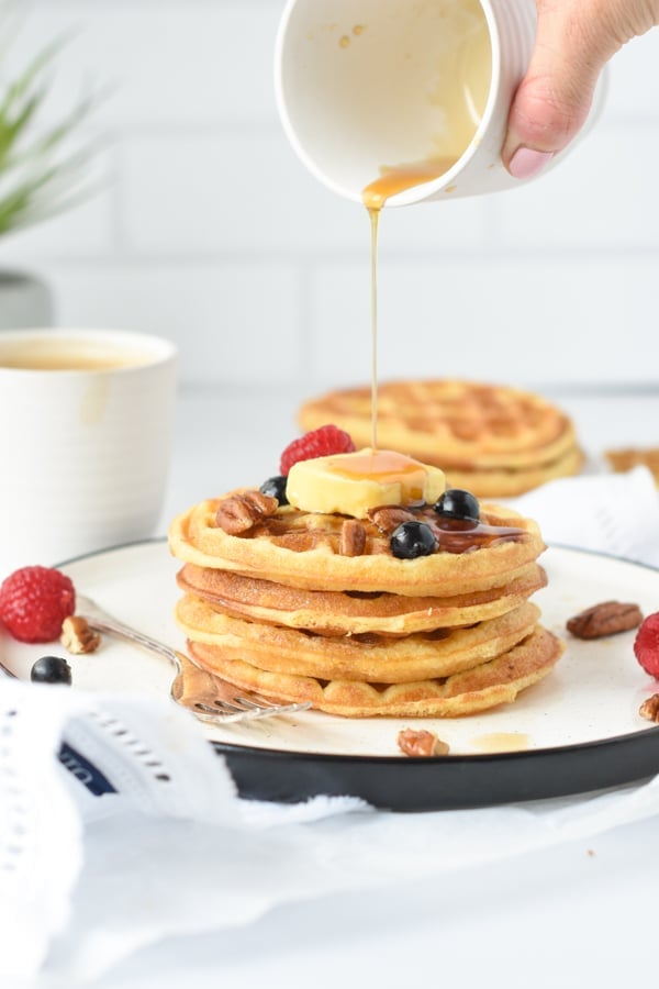 Pouring sugar-free maple syrup on top of a stack of coconut flour waffles decorated with butter, blueberries, and raspberries.
