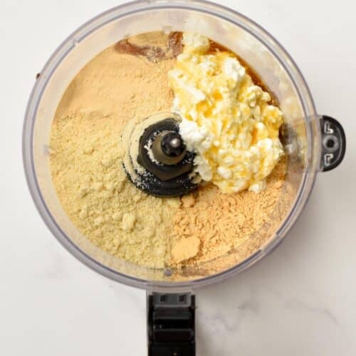 A food processor bowl filled with  almond flour, protein powder, powdered peanut butter, cottage cheese, maple syrup, and vanilla.