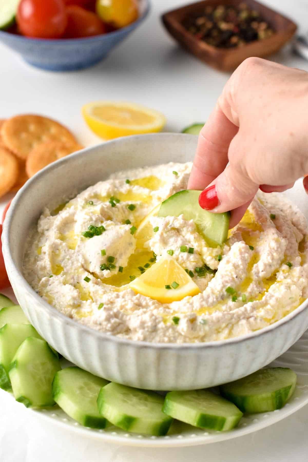 Dipping a slice of cucumber in Cottage Cheese Dip on a plate decorated with vegetables and crackers.