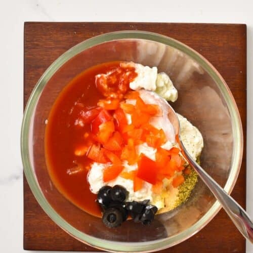 a large mixing bowl with cottage cheese, slices black olives, red bell pepper pieces, garlic powder, dried herbs and marinara sauce with a spoon in the bowl