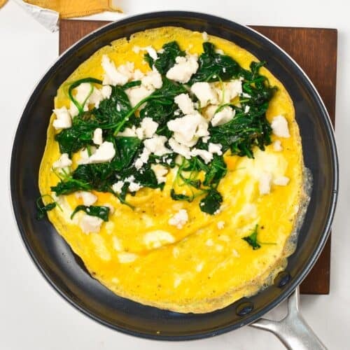 a feta omelette in a skillet, unfolded and filled on half of its side with feta cheese and cooked spinach