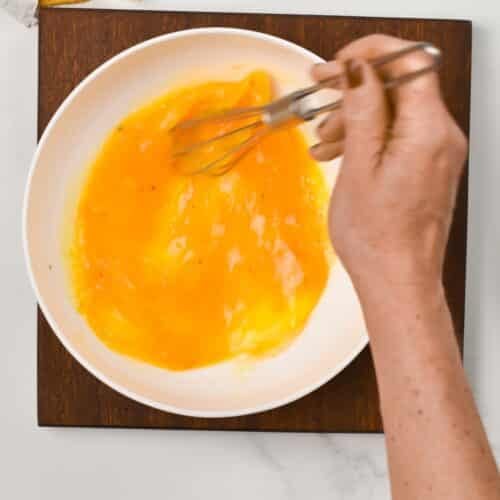 a hand beating eggs in a bowl with a small whisk