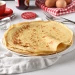 A Stack of French Crepe on a white tablecloth in front of a basket with eggs and a pot of jam.