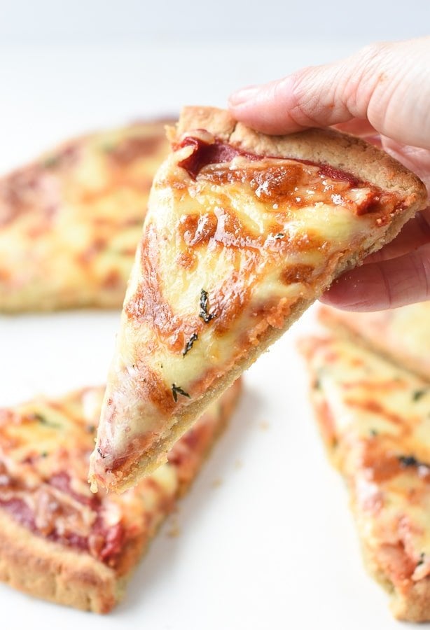 A hand holding a slice of Almond Flour Pizza Crust topped with cheese and tomato paste.