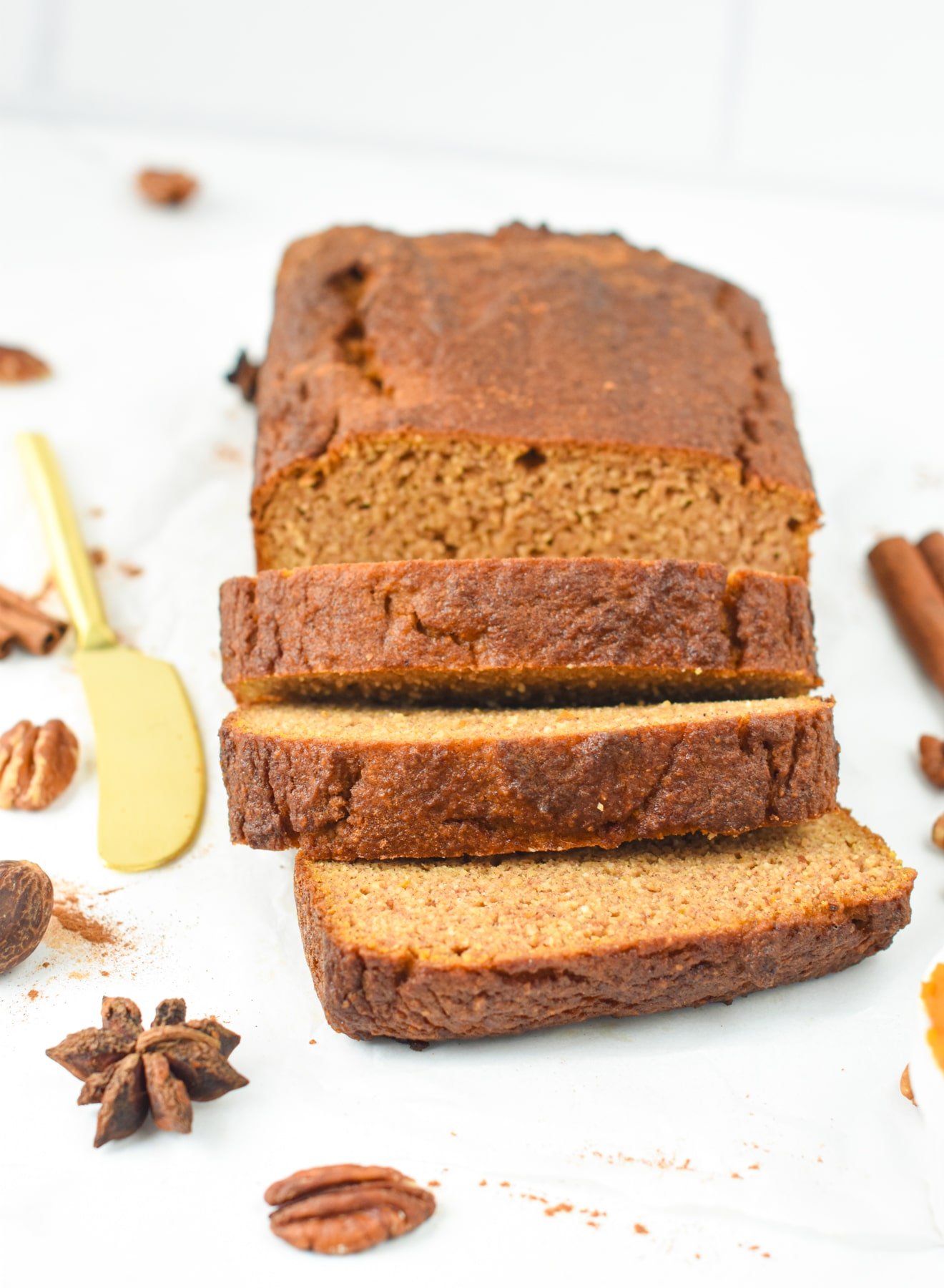 A loaf of Almond Flour Pumpkin Bread with 3 slices, next to a golden butter knife and spices.