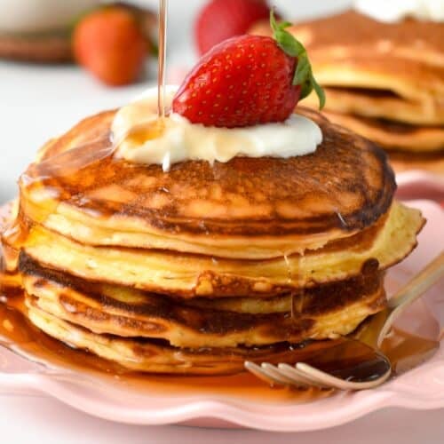 a stack of cottage cheese pancakes with a drizzle of maple syrup poured on top. The stack is also topped with yogurt presented on a pink plate, with a golden fork on side