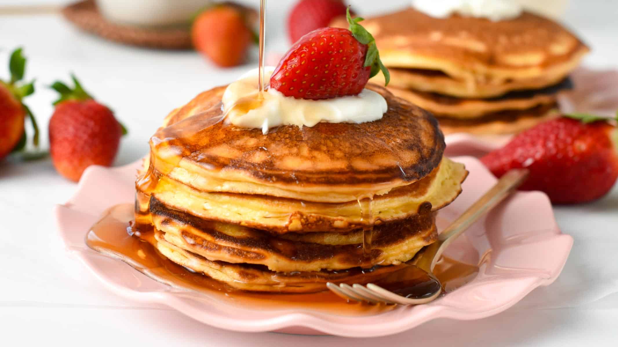 A stack of cottage cheese pancakes with a drizzle of maple syrup poured on top. The stack is also topped with yogurt presented on a pink plate, with a golden fork on side.