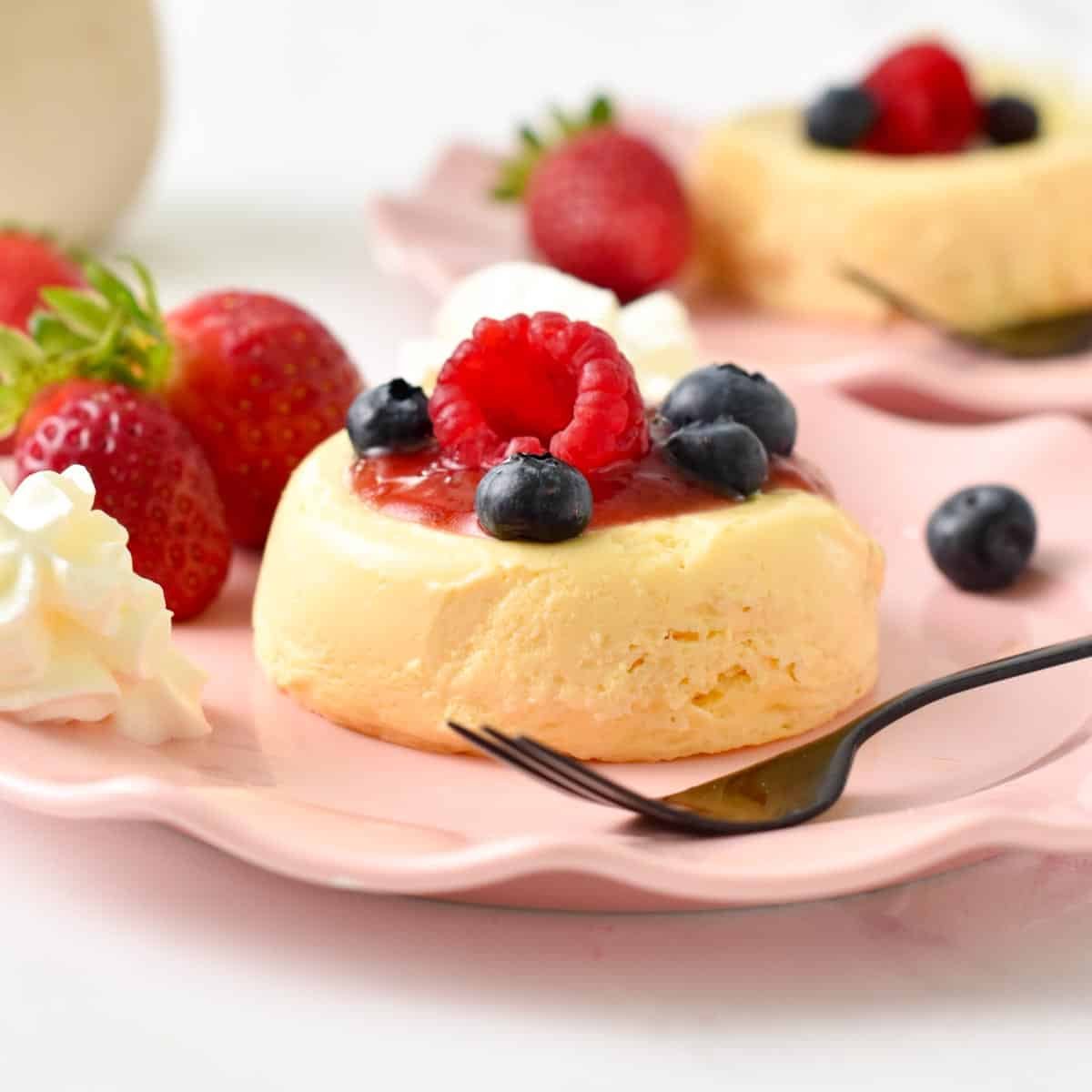 a single serve microwave cheesecake on a pink plate topped with jam, raspberry, blueberries and strawberries in the background, and a black fork in the front