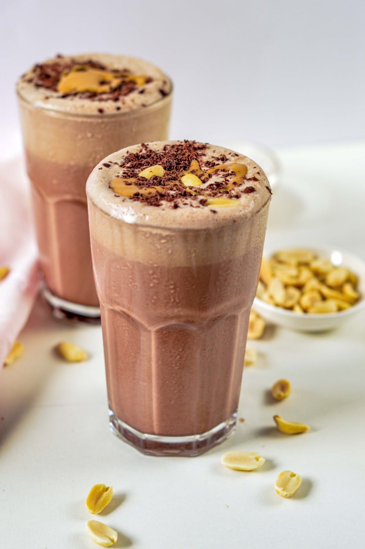 Keto Peanut Butter Smoothie decorated with chopped chocolate and fresh peanut butter in two large glasses surrounded with peanuts.