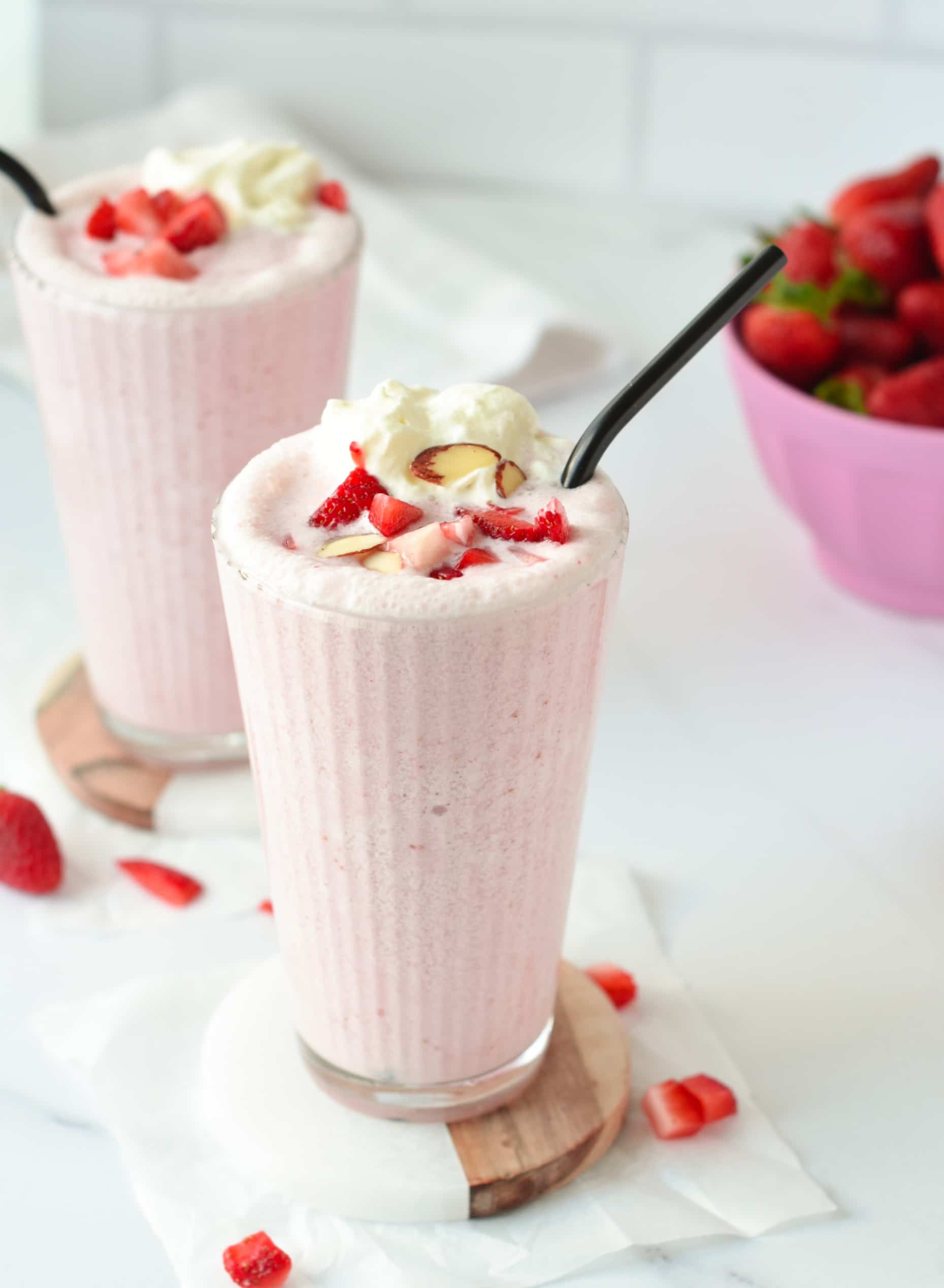 Two Keto Smoothie in tall glasses with black straws,  and decorated with whipped cream, chopped strawberries, and slivered almonds.
