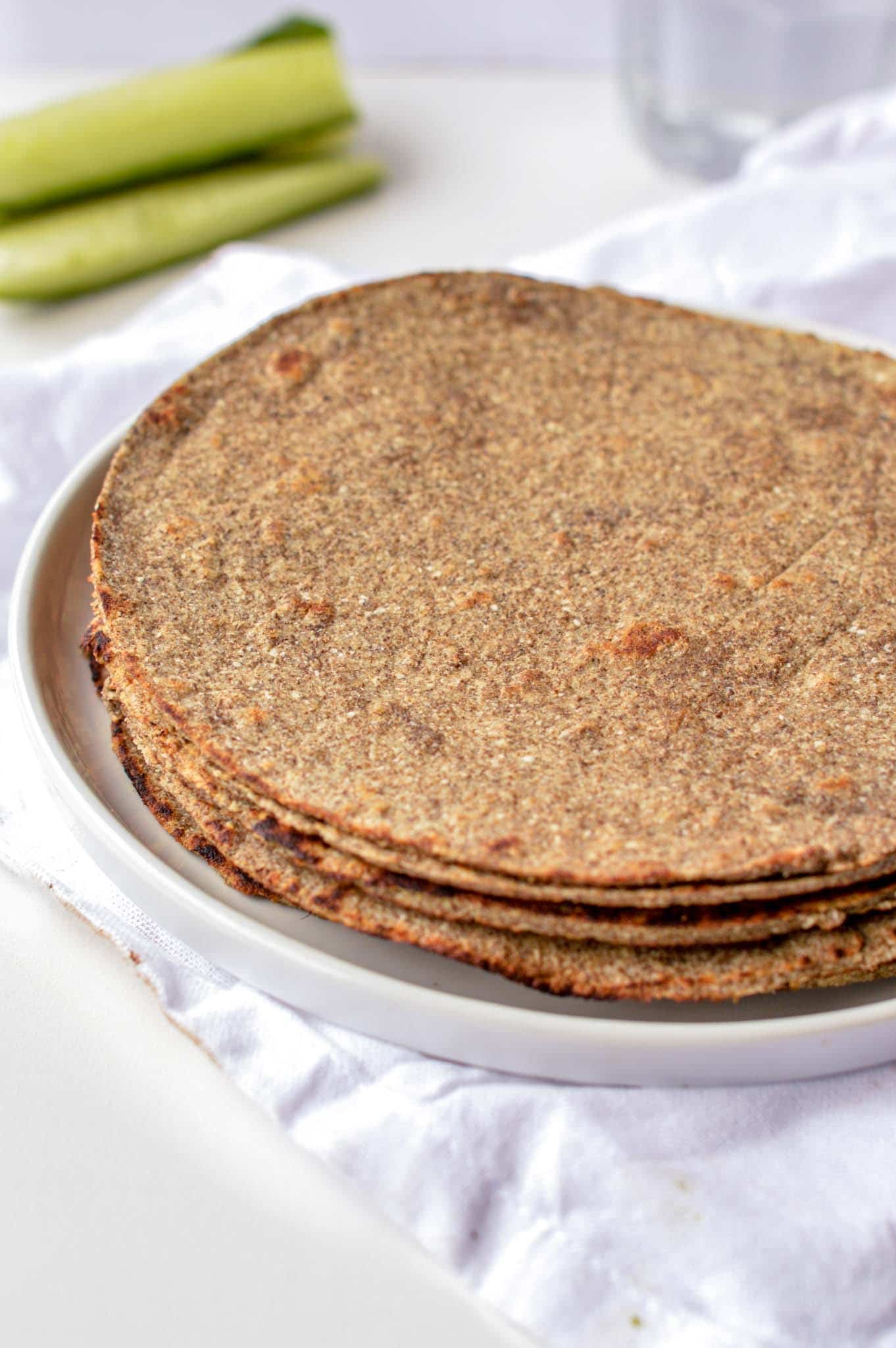 A stack of Keto Tortillas with Chia Seeds on a white plate with zucchinis in the background.