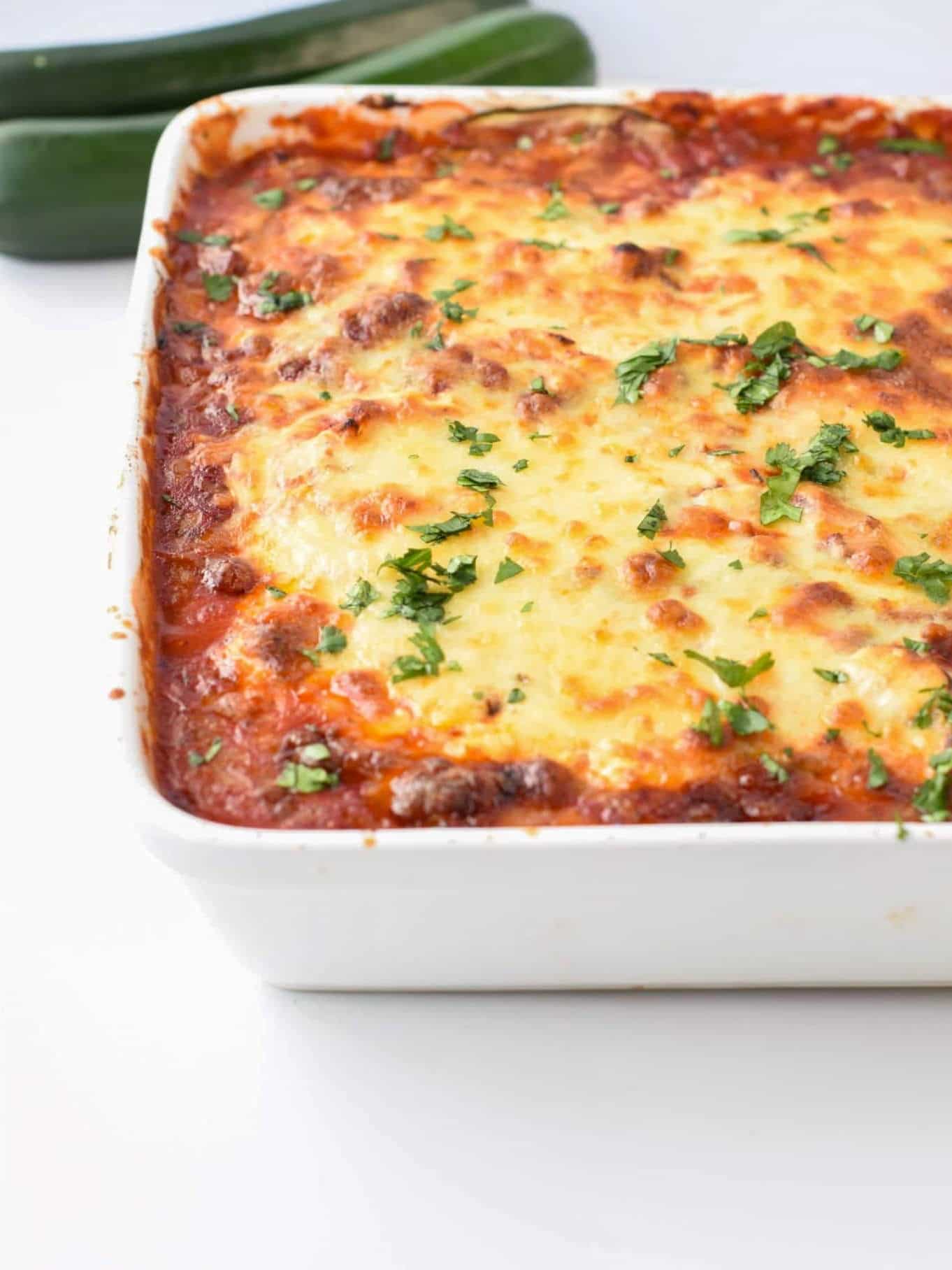Keto Zucchini Lasagna in their cooking pan in front of fresh zucchini.