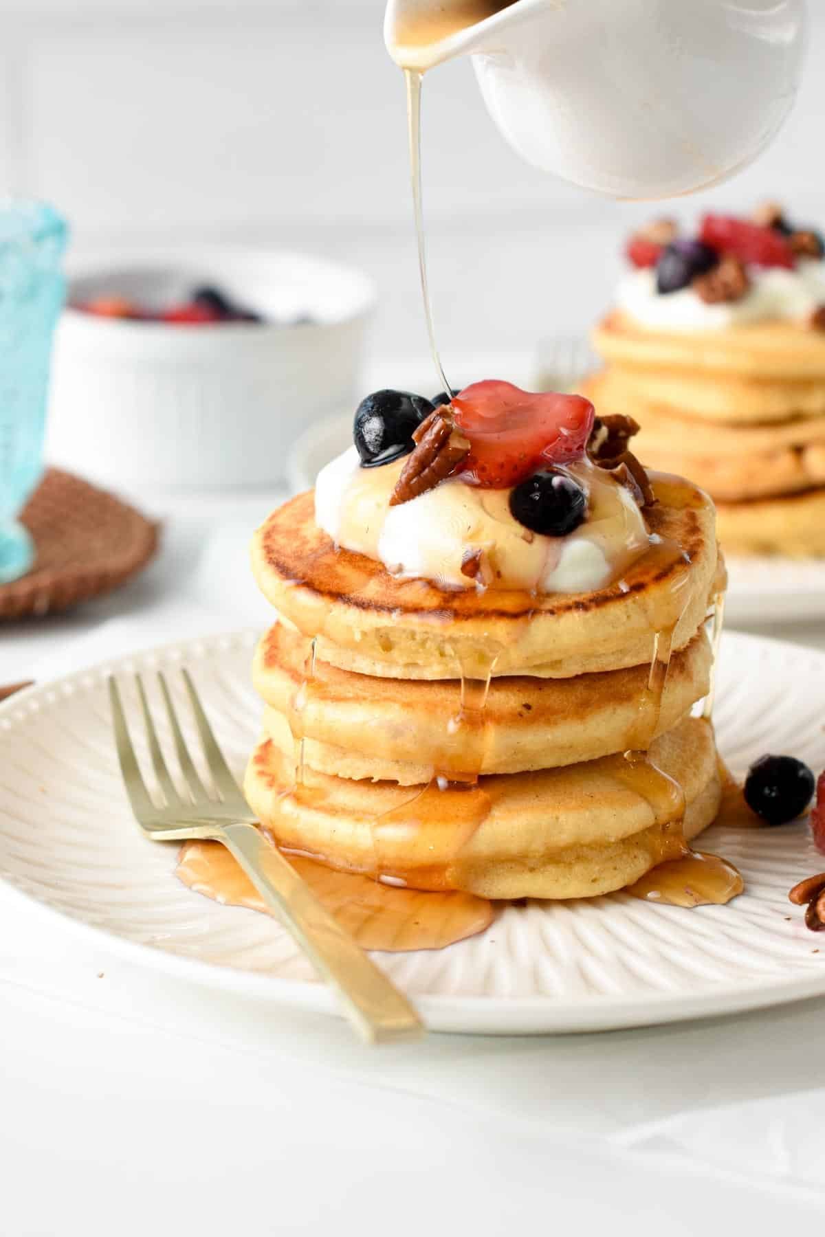 A stack of Low Carb Protein Pancakes decorated with coconut cream, strawberry slices, blueberries, pecan nuts, and a drizzle of maple syrup.