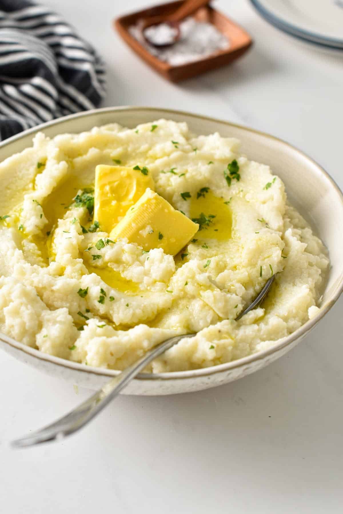 Mashed Cauliflower in a large bowl with a spoon and decorated with fresh herbs and garlic butter.