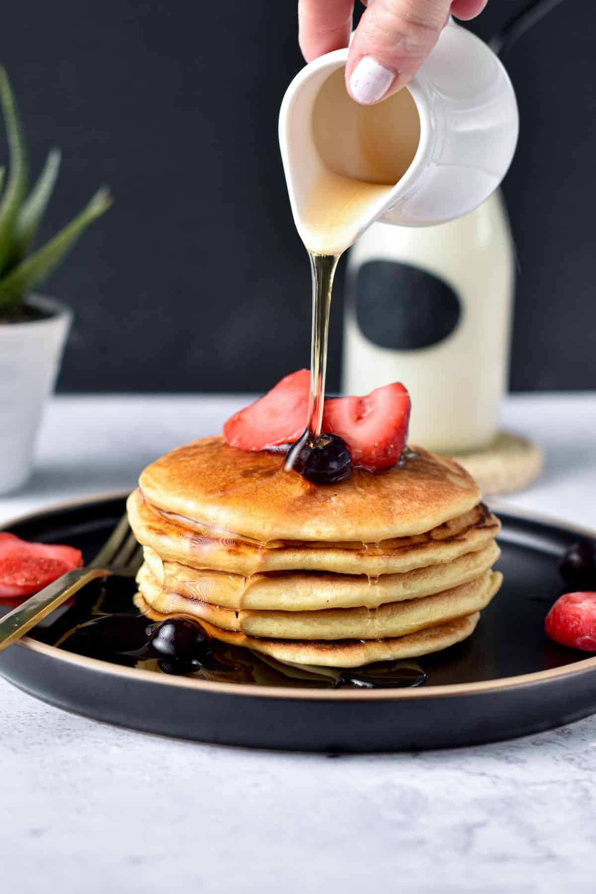 Pouring maple syrup on Paleo Pancakes decorated with strawberries and blueberries on a black plate.