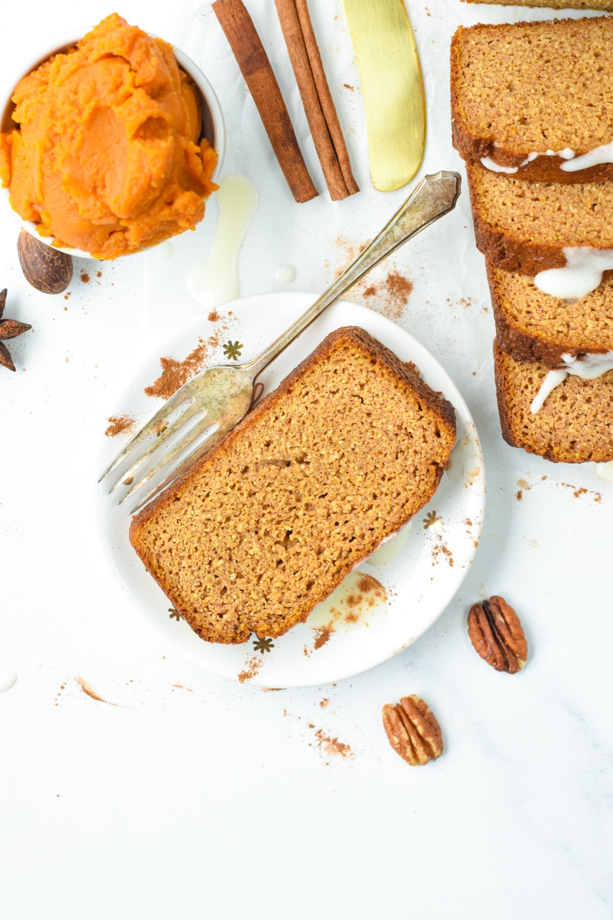 A slice of Pumpkin Bread with Almond Flour on a small white plate next to several other slices, cinnamon sticks, and pumpkin puree.,