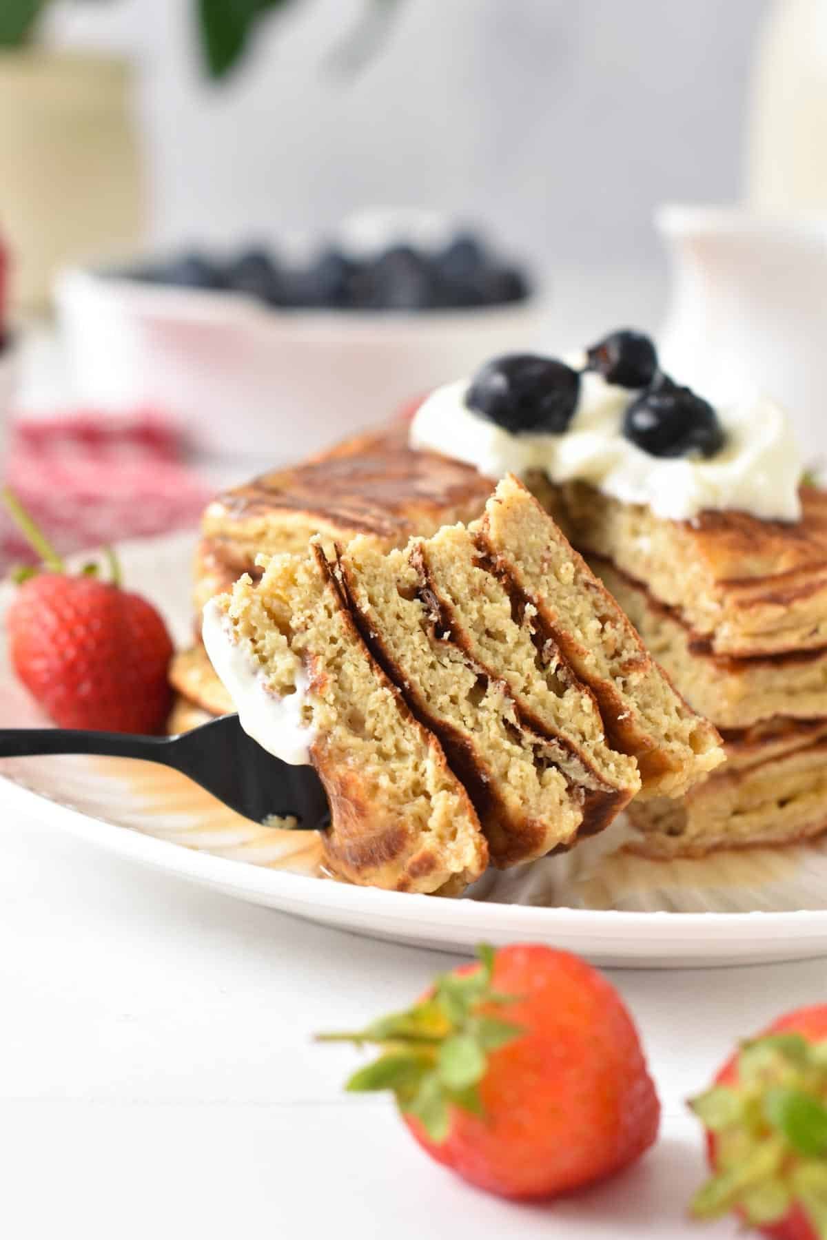 A stack of protein pancakes with Greek yogurt, blueberries on top and in the front, a fork with a pieces of cut pancakes showing their fluffy texture.