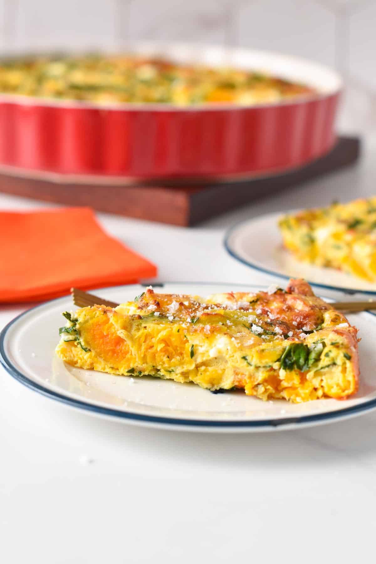 A slice of pumpkin frittata in a plate with the full frittata in the background.