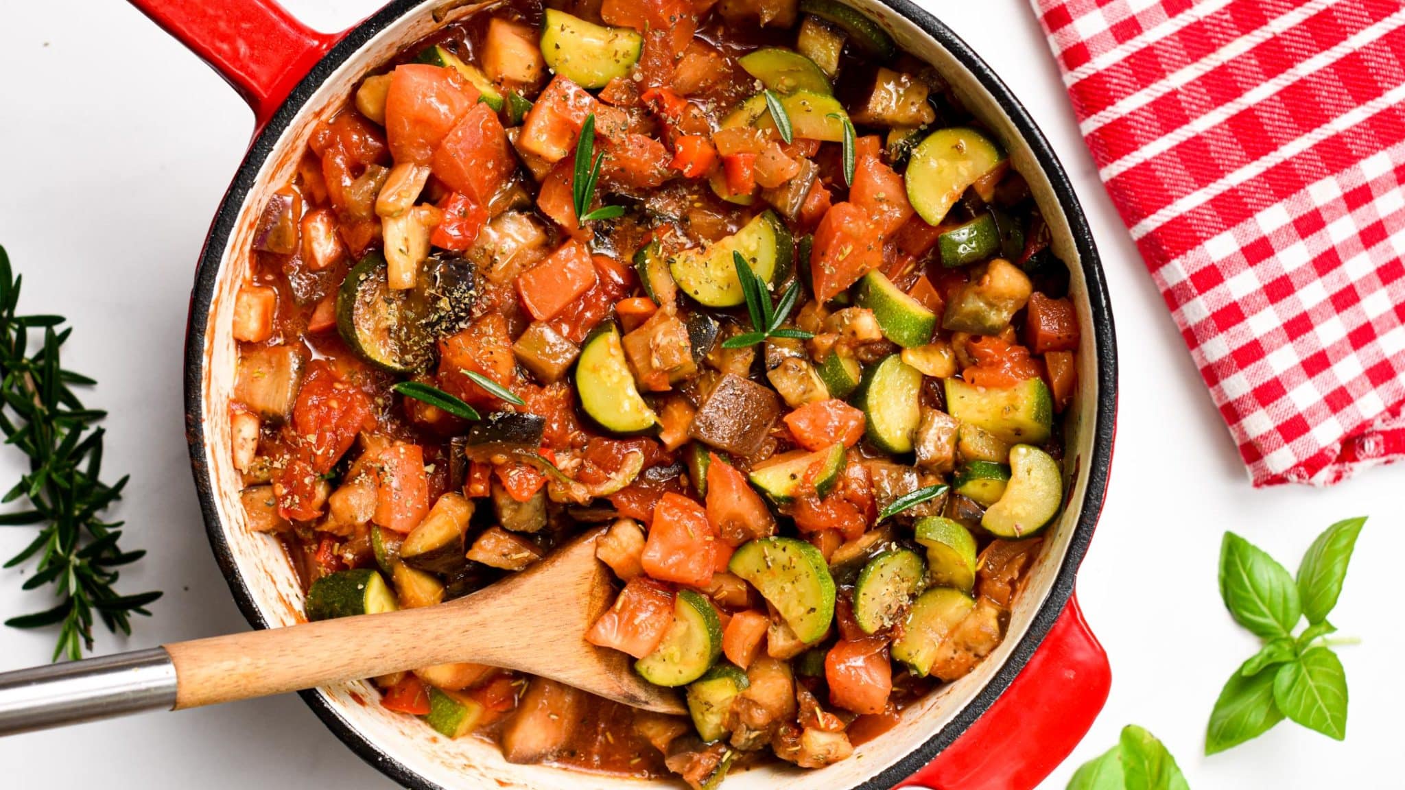 Ratatouille recipe in a large cast-iron Dutch oven with a wooden spoon with basil and rosemary next to it.