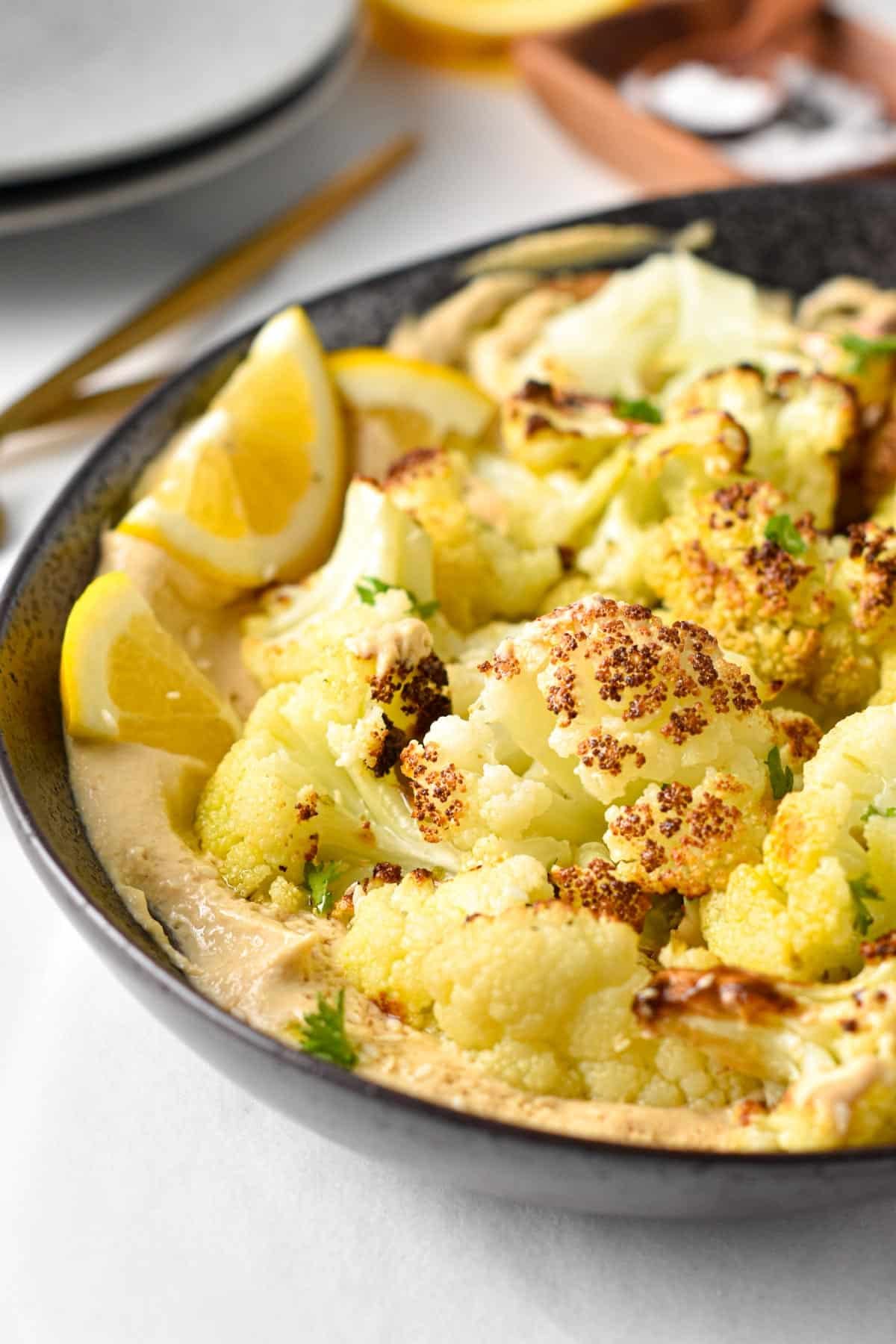 A dish of roasted cauliflower florets on top of a creamy tahini sauce with lemon wedges.