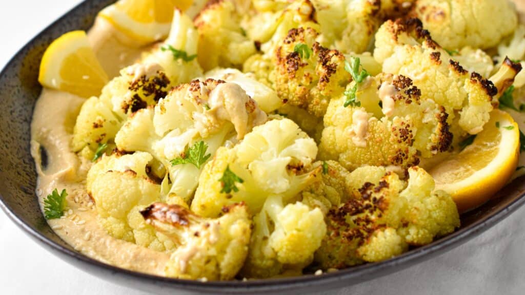 a dish of roasted cauliflower florets on top of a creamy tahini sauce with lemon wedges