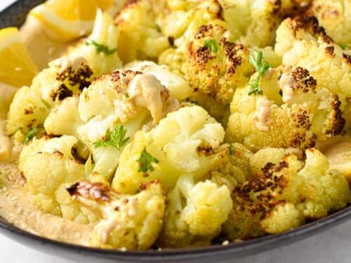 a dish of roasted cauliflower florets on top of a creamy tahini sauce with lemon wedges