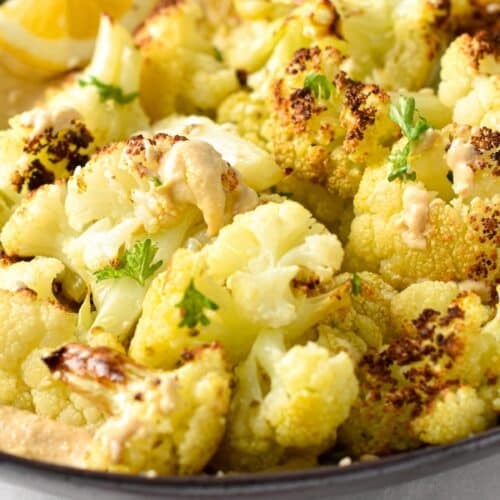 a dish of roasted cauliflower florets on top of a creamy tahini sauce with lemon wedges