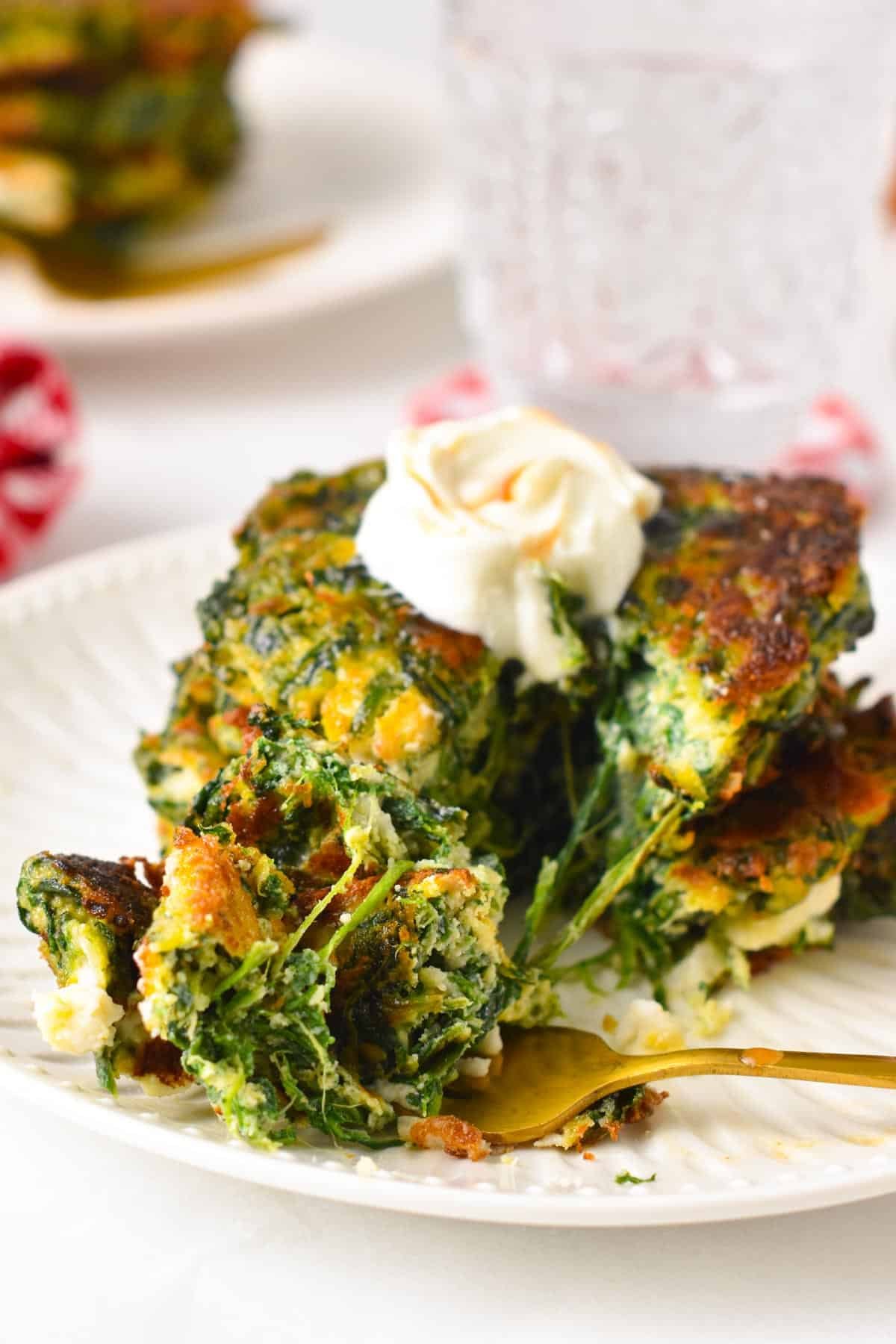 a stack of spinach fritters on a white plate with a golden fork cutting the fritters and showing the inside texture