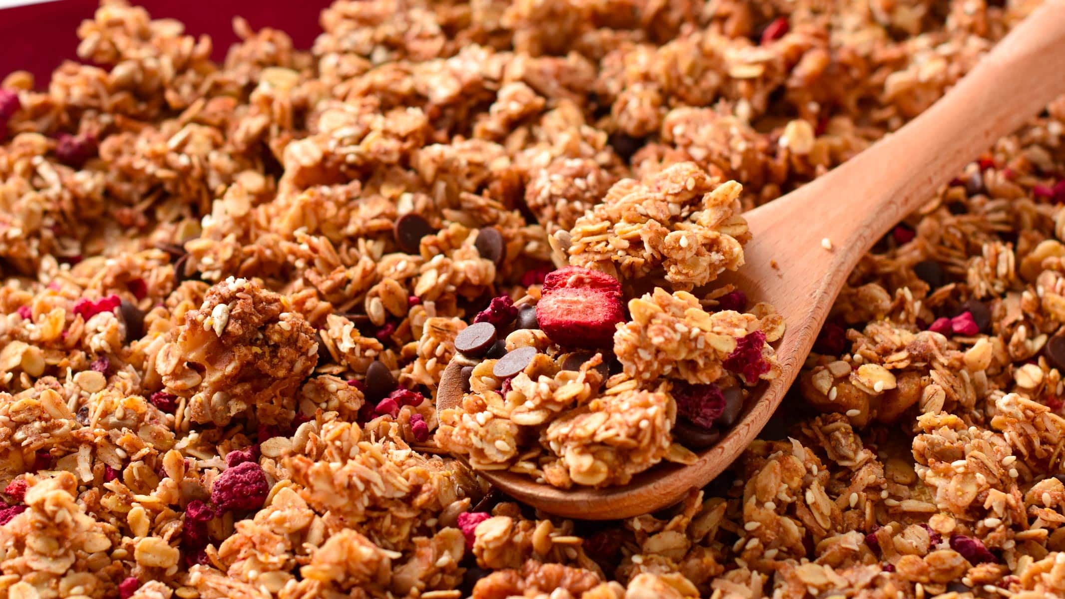 A baking sheet with a large batch of baked sugar-free granola and a wooden spoon with a serving.