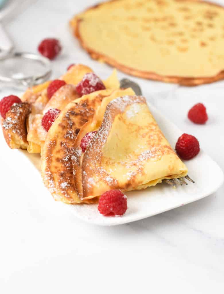 Keto Crepes folded on a white plate and decorated with powdered sweetener and fresh raspberries.