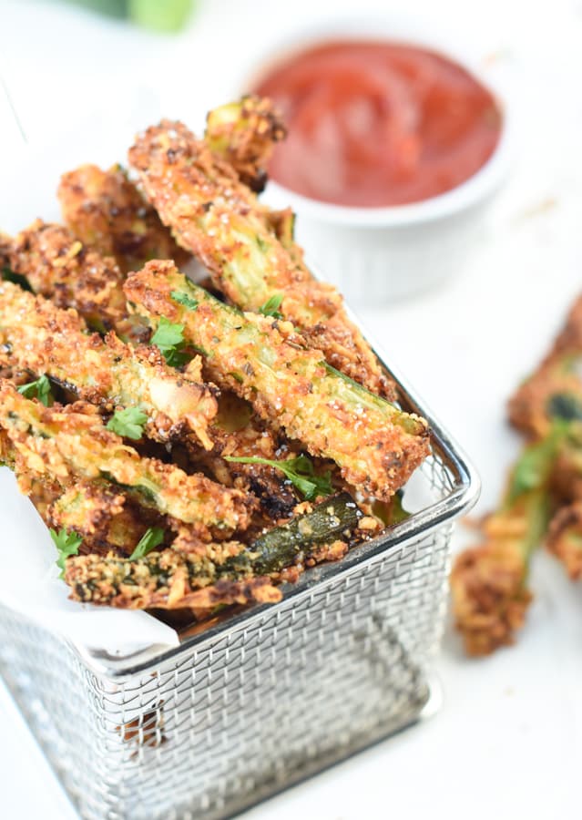 Keto Zucchini Fries with Almond flour on a small deep frying basket next to a jar with tomato sauce. 