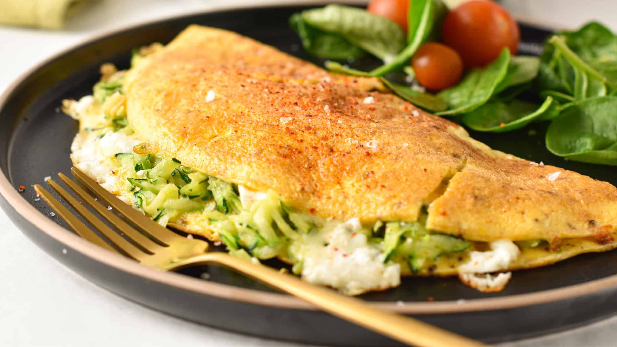 A zucchini omelette filled with feta and Parmesan on a black plate decorated with baby spinach and tomatoes.
